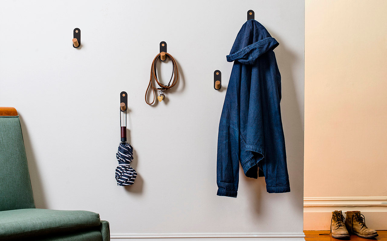 A wall hook with wooden pegs, the Plane Single Hook - Set of 3 by Fire Road, holds a coat, belts, and a striped scarf. A pair of shoes is on the floor adjacent to the wall's base. The edge of a teal sofa is visible in the foreground.
