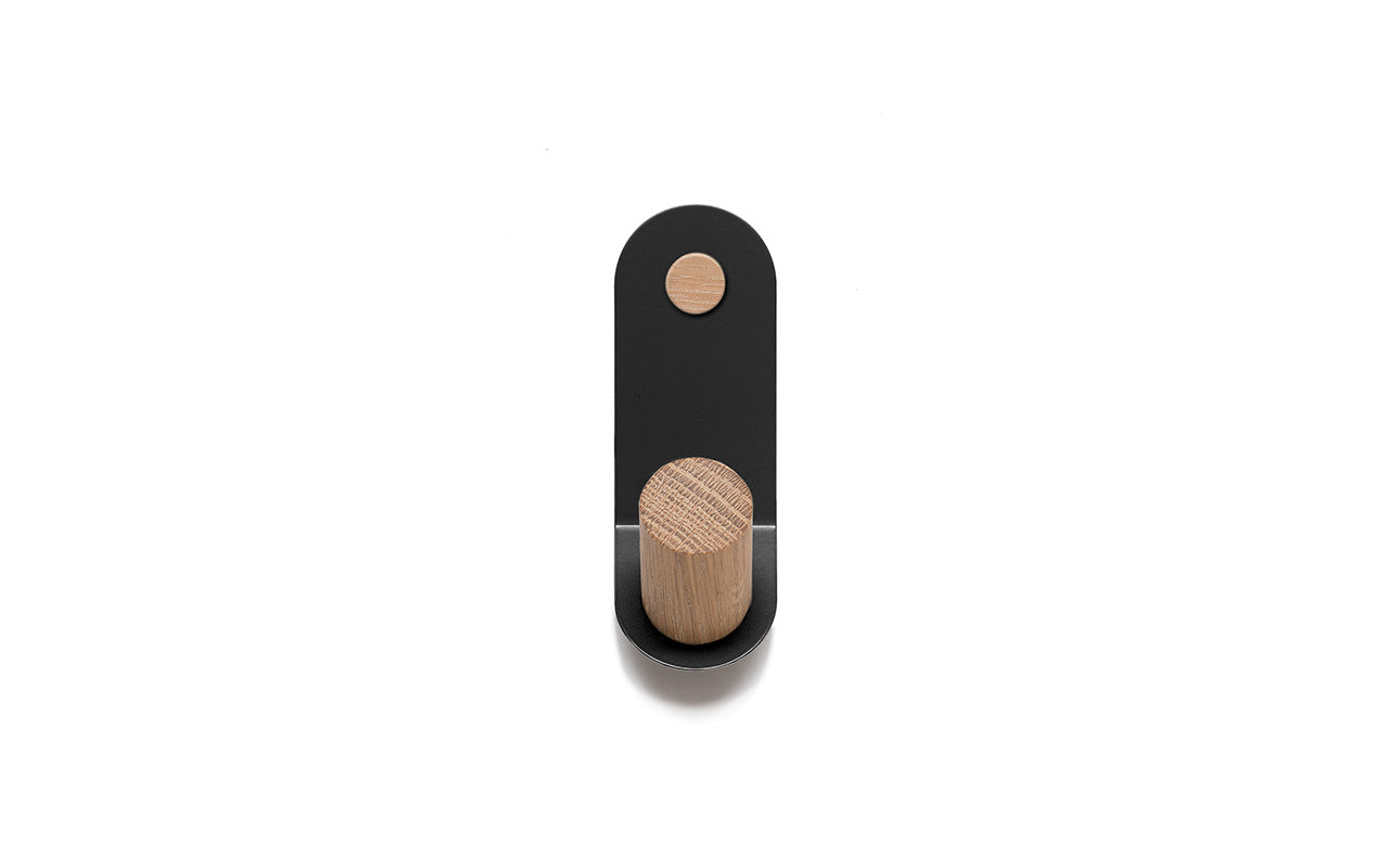 The Plane Single Wall Hook by Fire Road showcases a minimalist design with a black, powder-coated steel plate that includes a circular cutout and a cylindrical wooden peg.