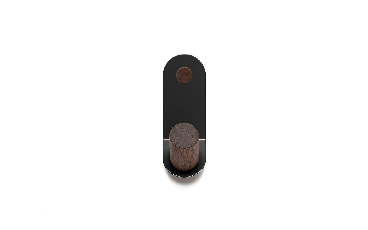 The Plane Single Wall Hook by Fire Road is a minimalist black and wooden wall hook featuring an oblong base and a circular knob made of wooden peg, mounted on a white background.
