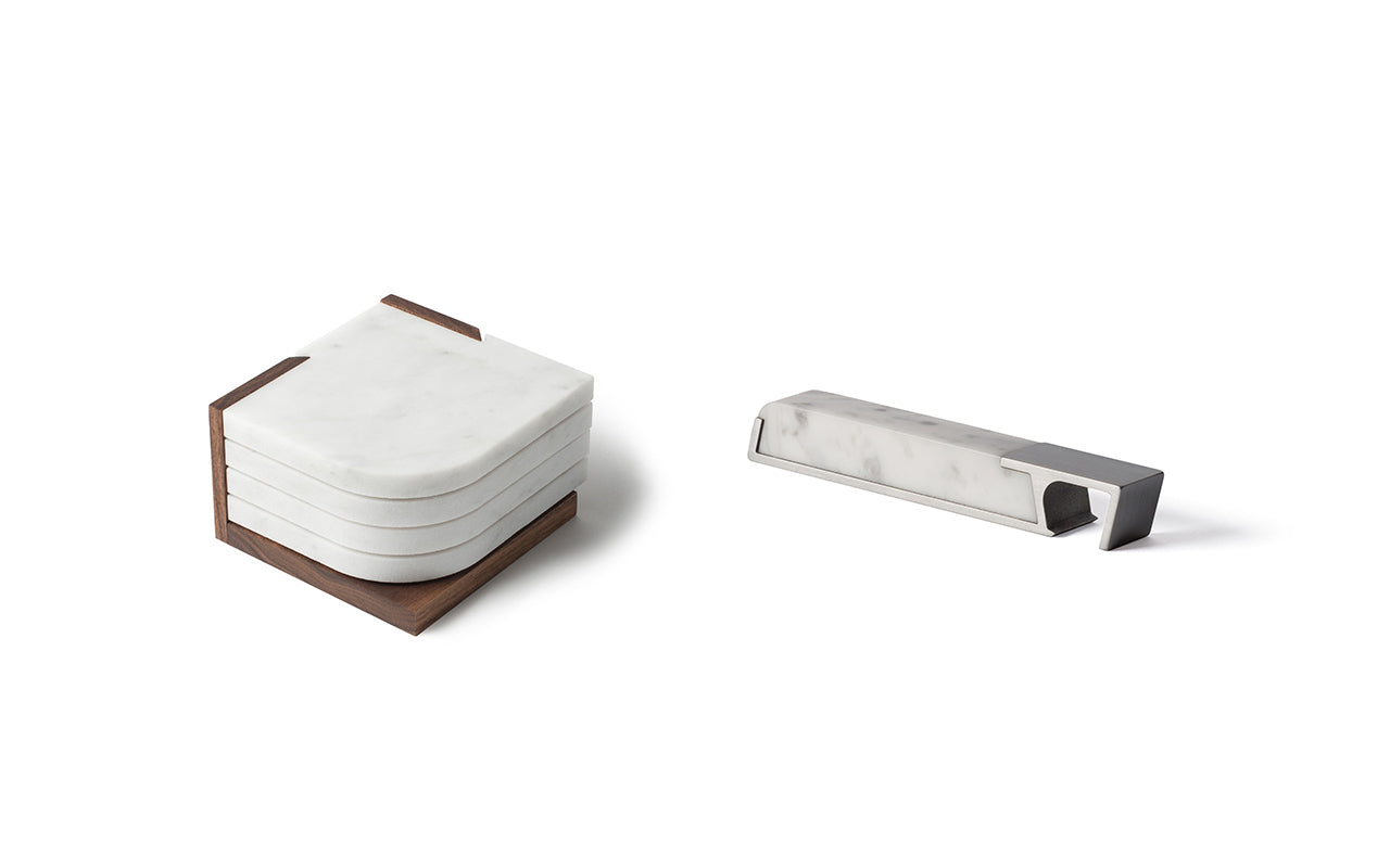 A set of four stacked, white marble coasters with a wooden holder on the left, and a sleek Profile Bottle Opener crafted from white marble and metal on the right, collectively known as the Fire Road Bar Set by Fire Road.