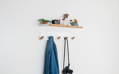 A white wall-mounted coat rack with five wooden hooks, shown with a blue denim jacket and a black camera strap hanging from it. Above the rack is a matching white shelf with small decorative plants and vases against a plain white wall.