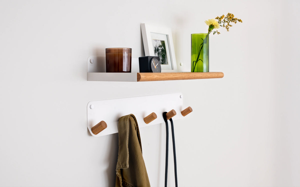 A minimalist wall-mounted storage set featuring a white shelf with a light wood accent and a matching white coat rack with wooden hooks, styled with home decor accessories against a clean white wall.
