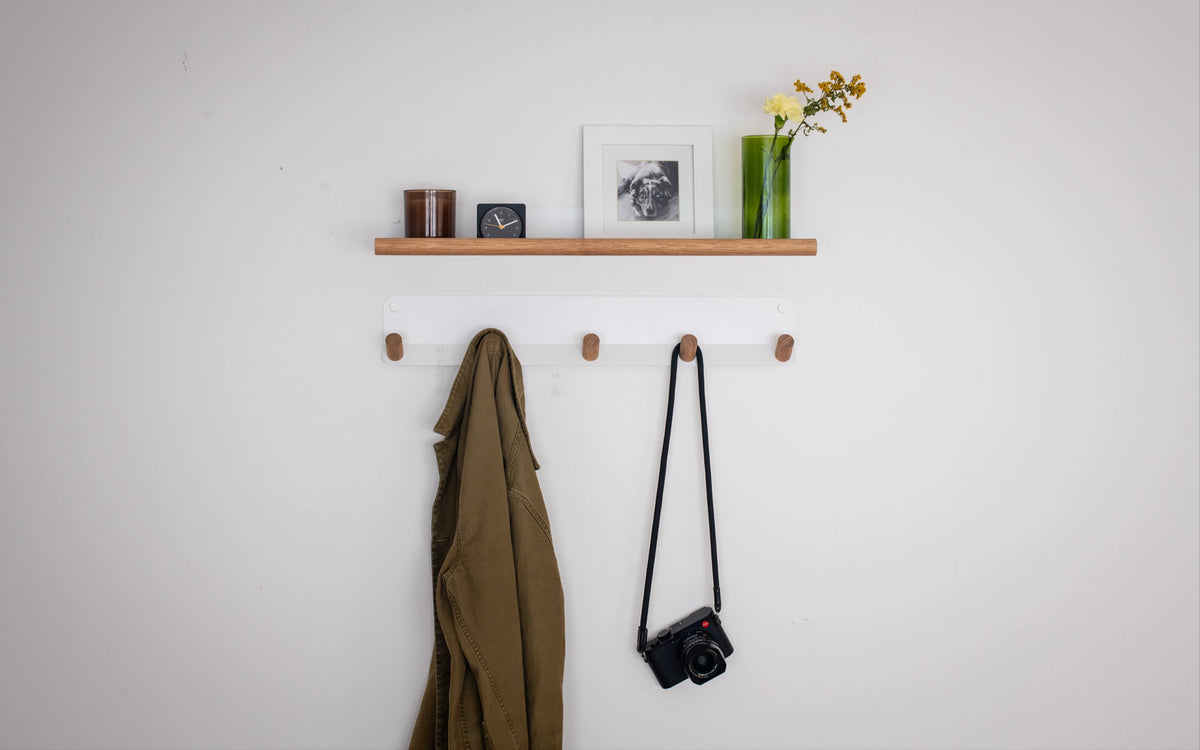 A minimalist wall-mounted wooden shelf and coat rack set on a white wall, styled with a green vase of flowers, a framed print, and small decor items. A brown jacket hangs on the lower rack hooks, and a black camera with a strap is also hanging.