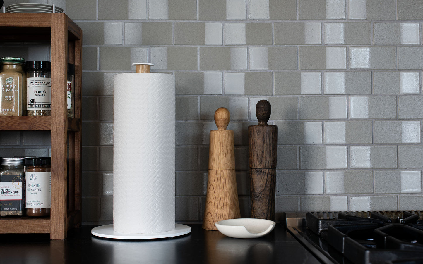 A countertop with a powder coated steel Plane Paper Towel Holder by Fire Road, two wooden pepper mills, a small dish, and a spice rack against a tiled backsplash. Made in the USA.