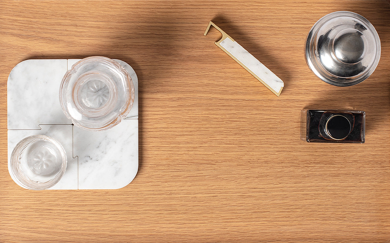 Wooden surface with marble coasters, two glass cups, a Fire Road Bar Set with a white handle, and a glass bottle with a dark cap by Fire Road.